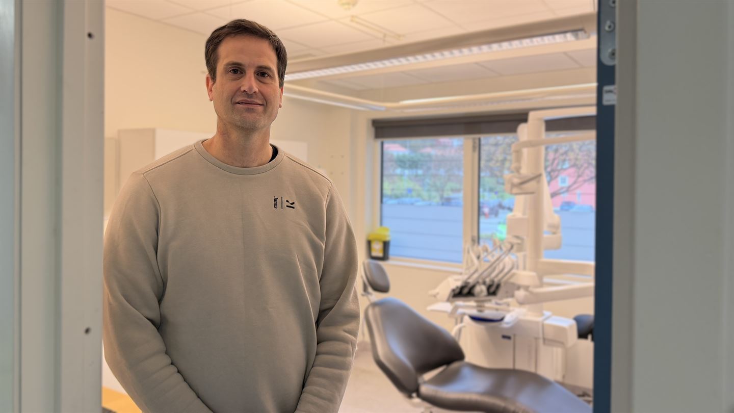 En mann står i en døråpning og vi ser inn i et rom med en tannlegestol.